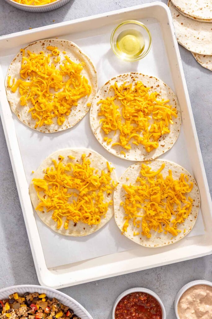 Shredded cheddar cheese sprinkled over tortillas for sheet pan quesadillas