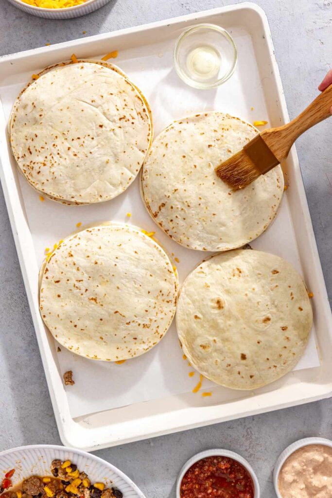 Second tortillas placed over the filling to assemble sheet pan quesadillas and brushing oil over top