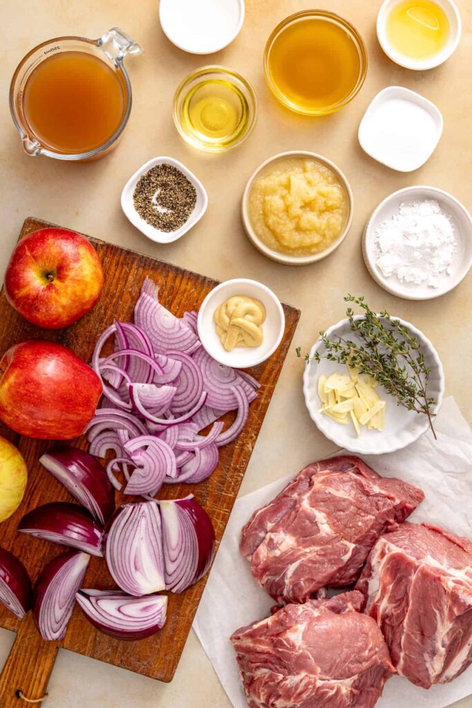 Ingredients for slow cooker apple cider pork, including pork shoulder, apples, red onion, garlic, fresh thyme, apple cider, applesauce, and broth.