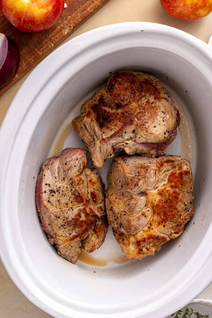 Seared pork shoulder pieces arranged in the slow cooker before adding the apple cider sauce.
