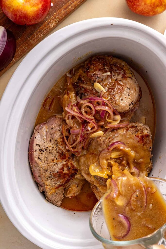 Apple cider sauce poured over seared pork shoulder in the slow cooker with fresh thyme sprigs.