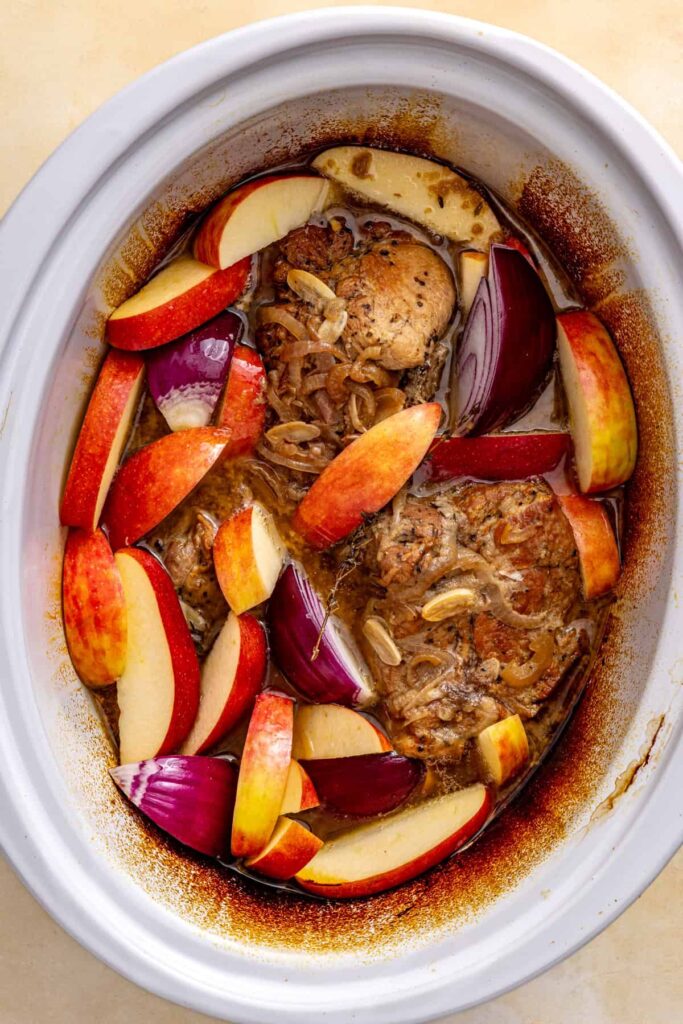 Thick apple wedges and red onion wedges nestled around pork shoulder in the slow cooker.