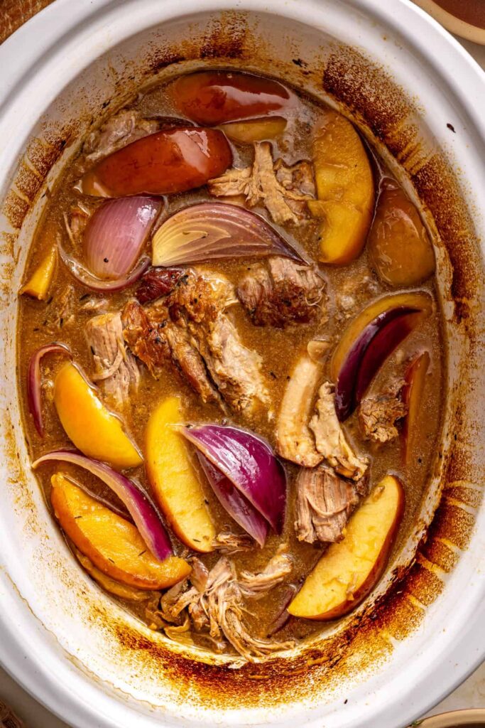Overhead view of slow cooker apple cider pork with shredded pork, apple wedges, red onion, and glossy cider gravy.