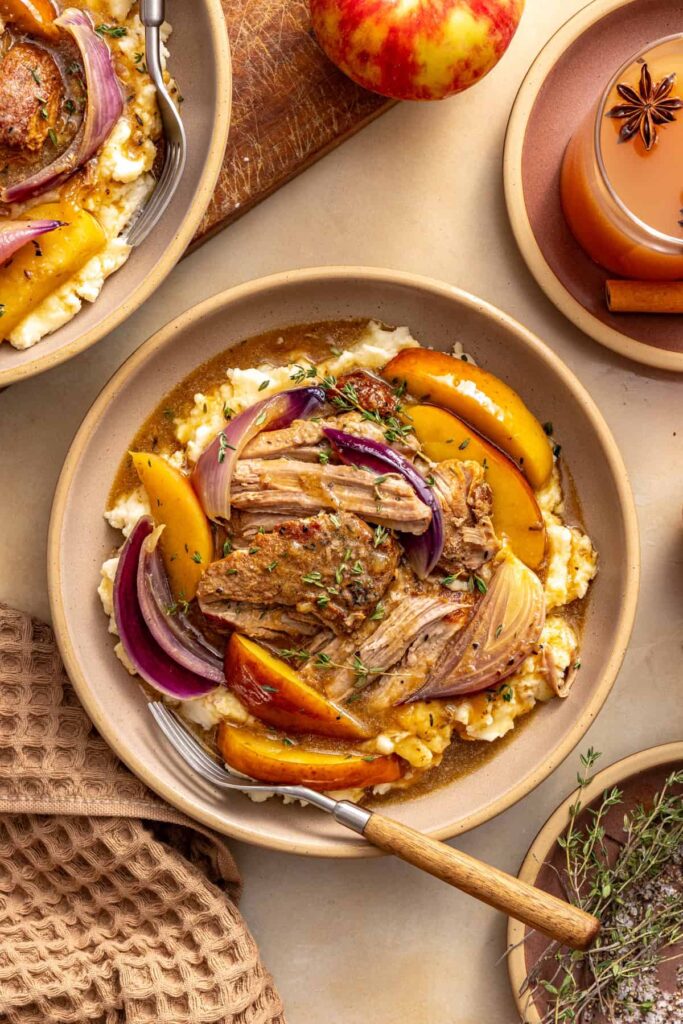 Slow cooker apple cider pork served over mashed potatoes with a fork, fresh thyme, and apples on a styled table.