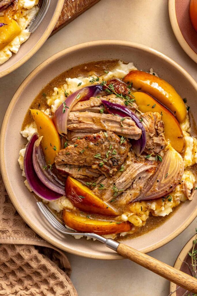 A bowl of slow cooker pork with apple cider gravy over mashed potatoes, styled with apples and a glass of cider.