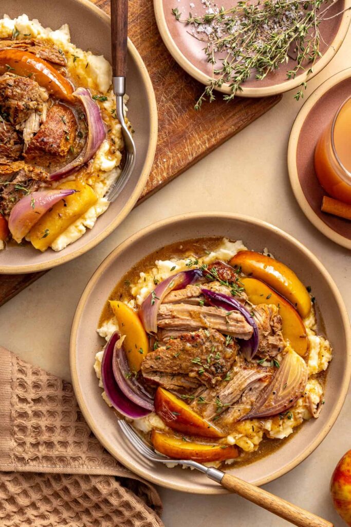 Two bowls of slow cooker apple cider pork served over mashed potatoes with fresh thyme and apples on a styled table.