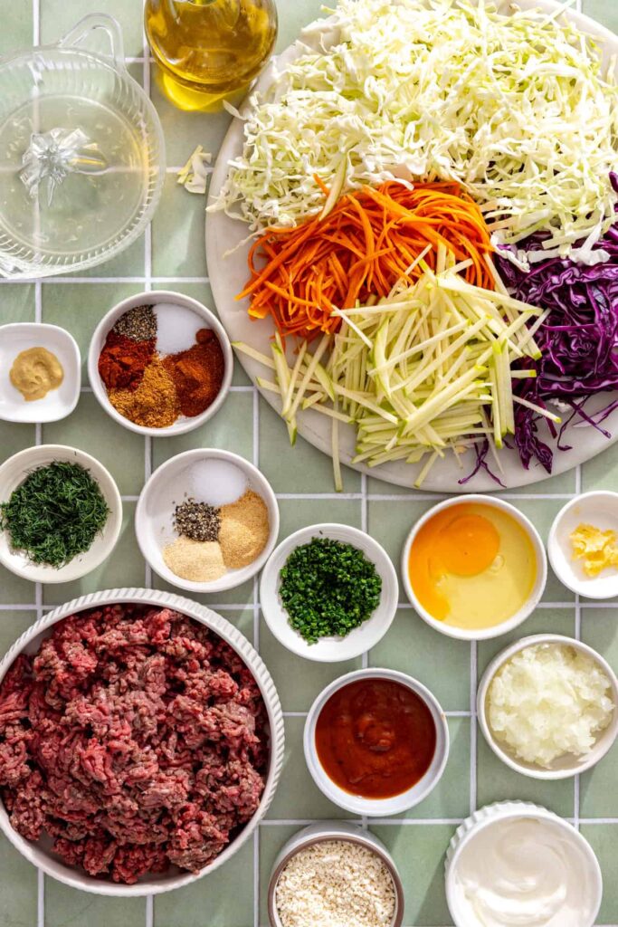 Overhead view of ingredients for BBQ meatballs and ranch slaw, including ground beef, breadcrumbs, cabbage, carrots, green apple, Greek yogurt, herbs, and BBQ sauce.