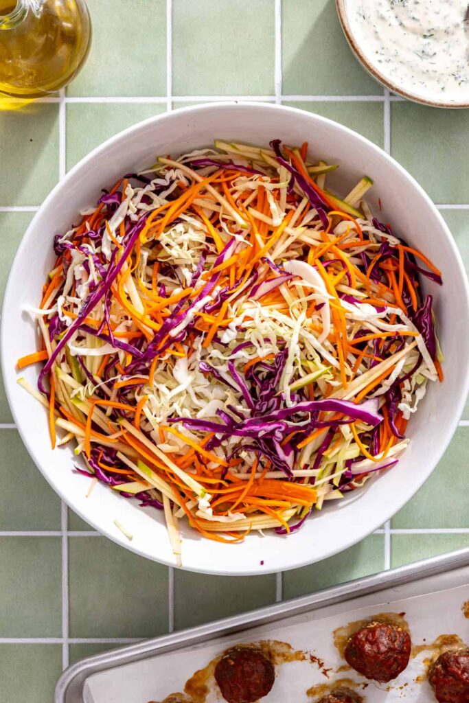 Shredded green cabbage, red cabbage, carrots, and green apple matchsticks tossed with fresh lemon juice in a large bowl.