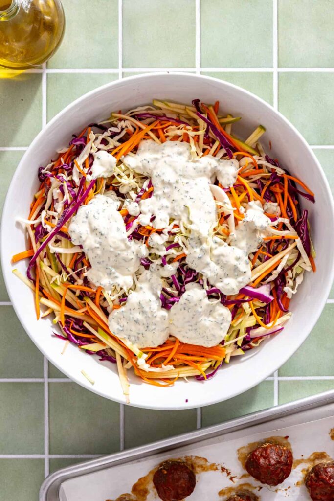 Pouring homemade Greek yogurt ranch dressing over shredded cabbage, carrots, and green apple for ranch slaw.