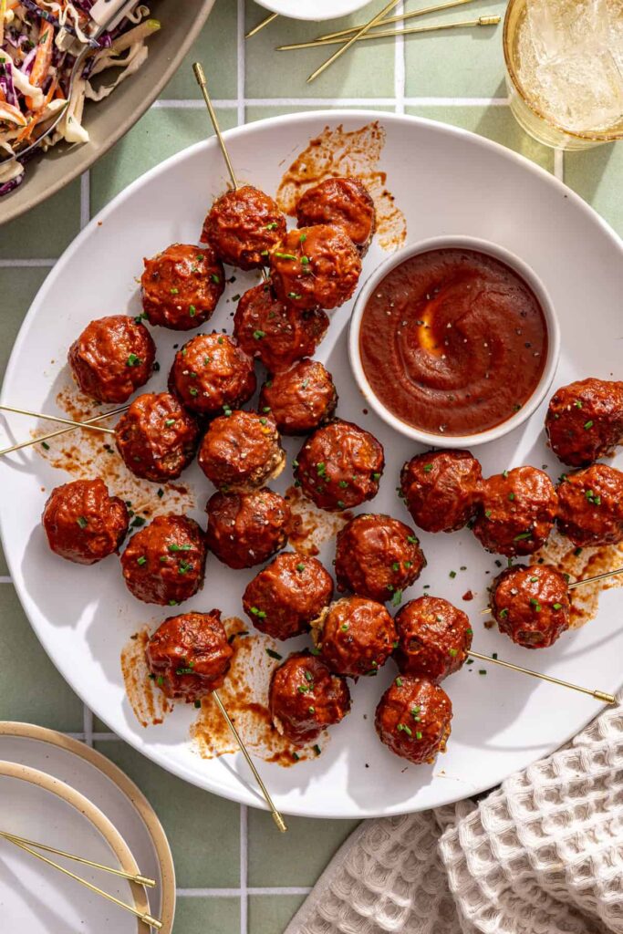 Overhead view of glazed BBQ beef meatballs on a plate with extra barbecue sauce and toothpicks for serving.