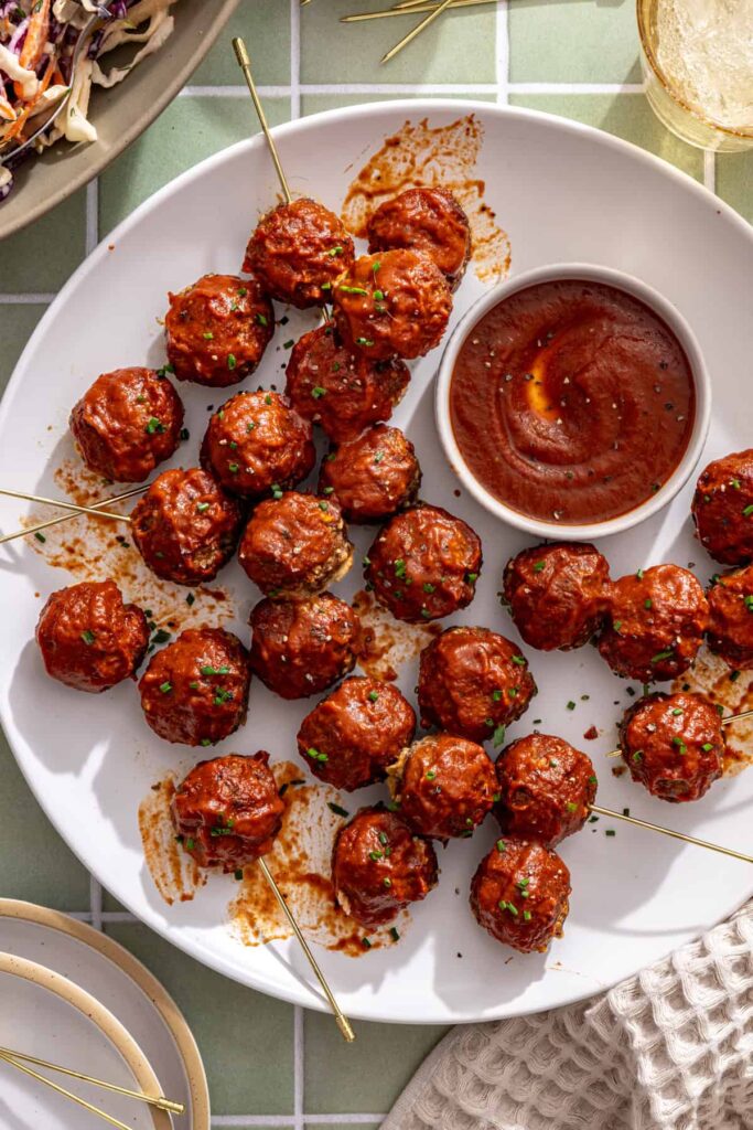 Homemade BBQ meatballs coated in sticky barbecue sauce, served with a small bowl of extra sauce.