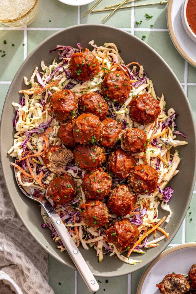 BBQ-glazed beef meatballs layered over colorful cabbage slaw with a serving spoon in the platter.