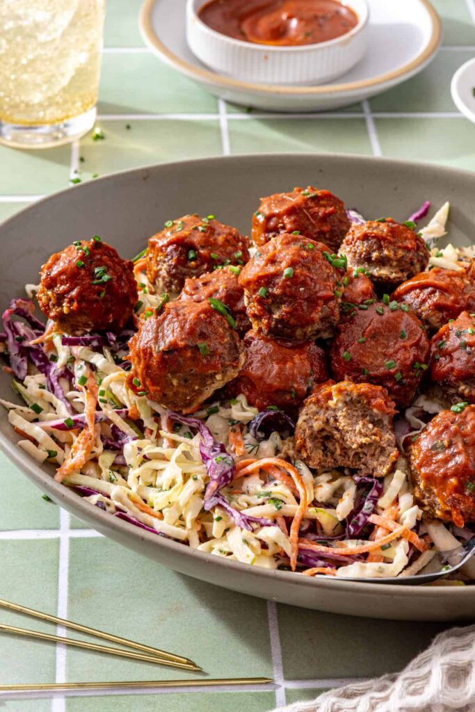 Side-angle close-up of glossy BBQ beef meatballs layered over creamy cabbage ranch slaw.