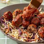 Close-up of BBQ meatballs glazed with barbecue sauce over creamy ranch slaw, with a half-eaten meatball showing its texture and a brush applying BBQ sauce.