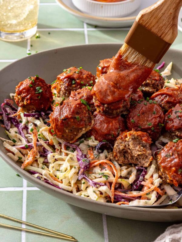 Close-up of BBQ meatballs glazed with barbecue sauce over creamy ranch slaw, with a half-eaten meatball showing its texture and a brush applying BBQ sauce.