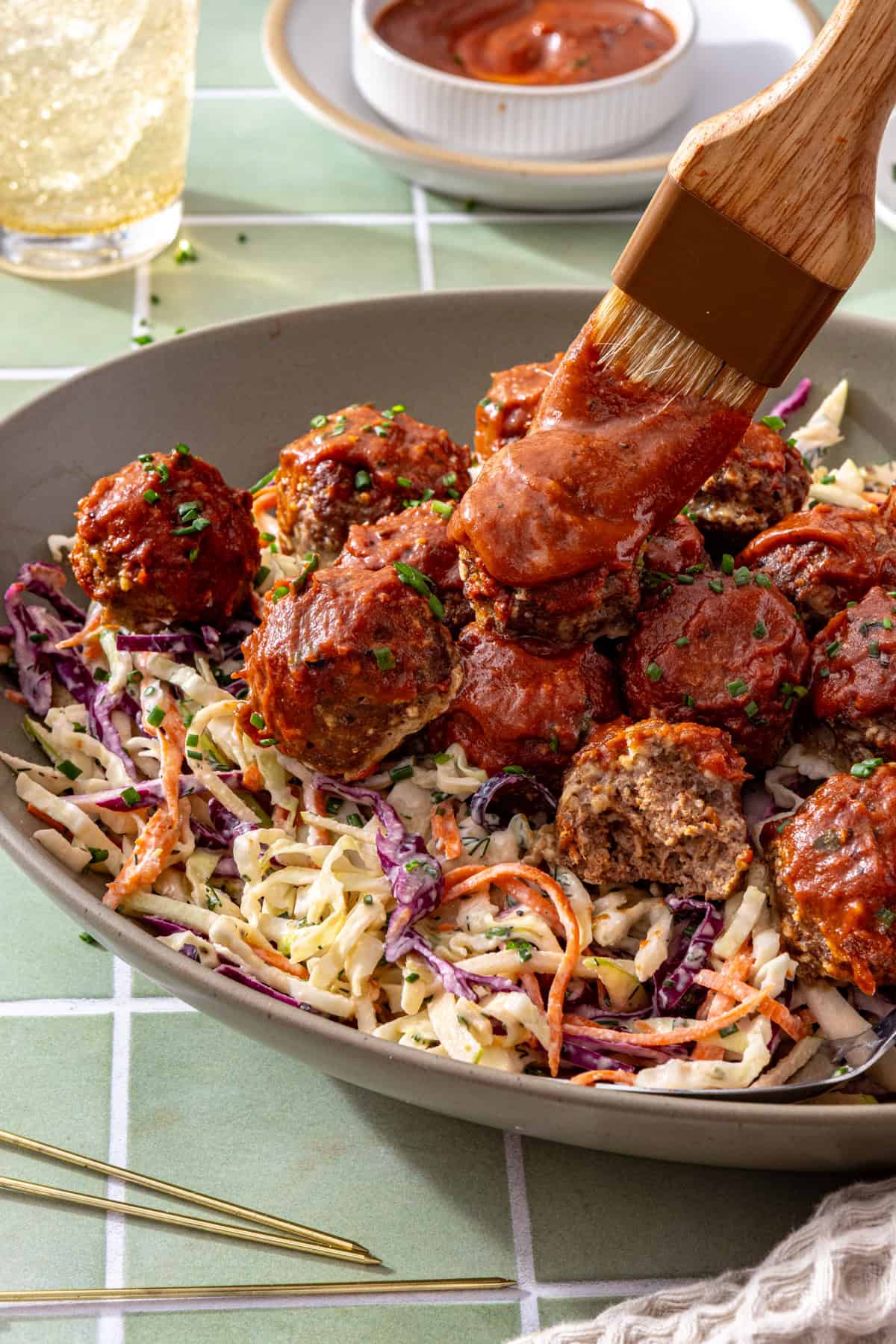 Close-up of BBQ meatballs glazed with barbecue sauce over creamy ranch slaw, with a half-eaten meatball showing its texture and a brush applying BBQ sauce.