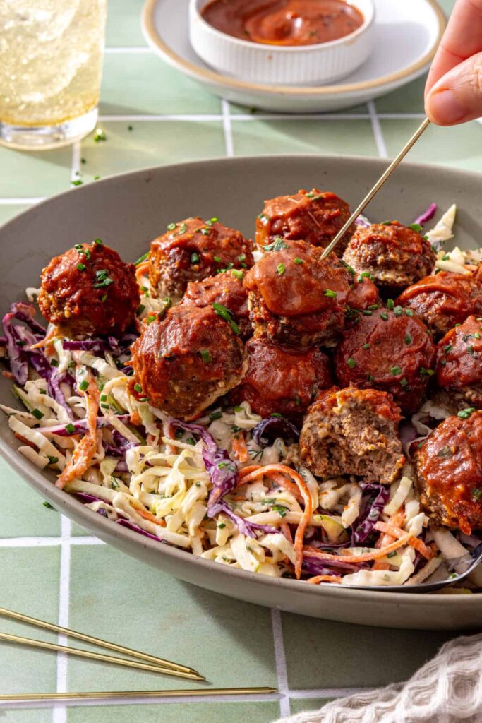Hand inserting a toothpick into a glazed BBQ meatball served over crunchy ranch slaw in a platter.