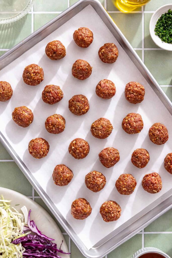 Raw homemade BBQ meatballs spaced evenly on a parchment-lined sheet pan before baking.