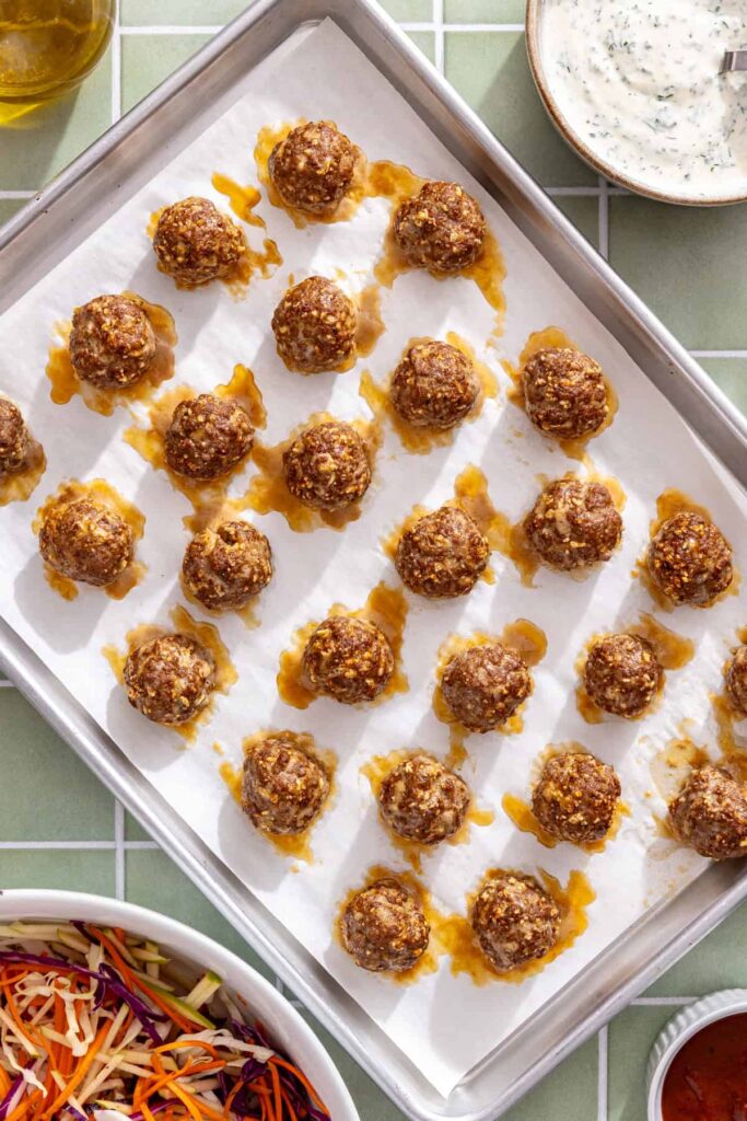 Partially baked homemade beef meatballs arranged on a baking sheet before adding BBQ sauce.