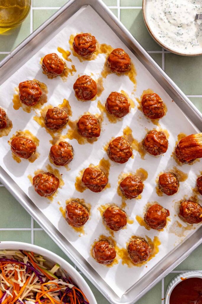Brushing BBQ sauce over oven-baked beef meatballs on a sheet pan.