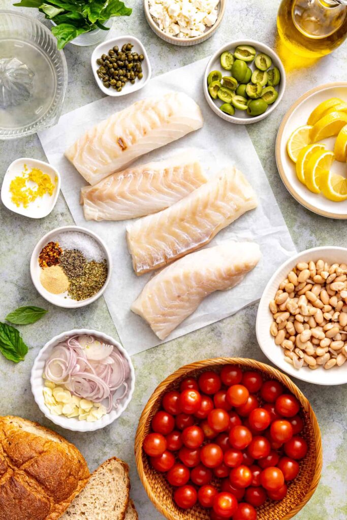 Overhead view of ingredients for Mediterranean baked cod, including cod fillets, cherry tomatoes, white beans, olives, feta, lemon, garlic, and fresh herbs.