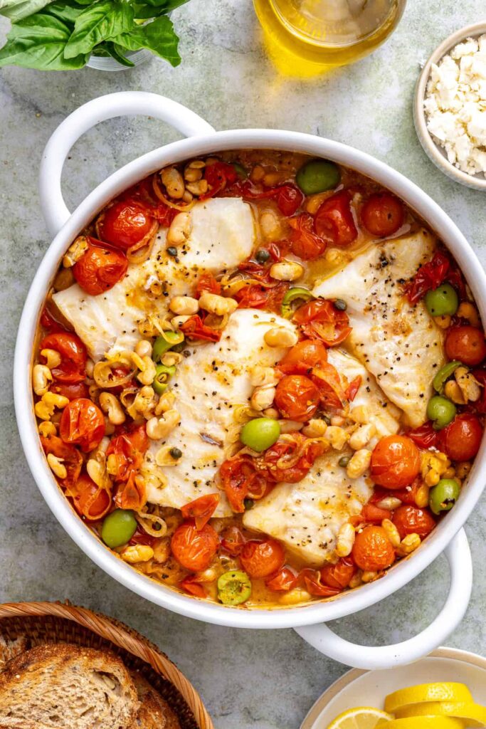 Cod fillets baking in a skillet with blistered tomatoes, cannellini beans, and green olives.