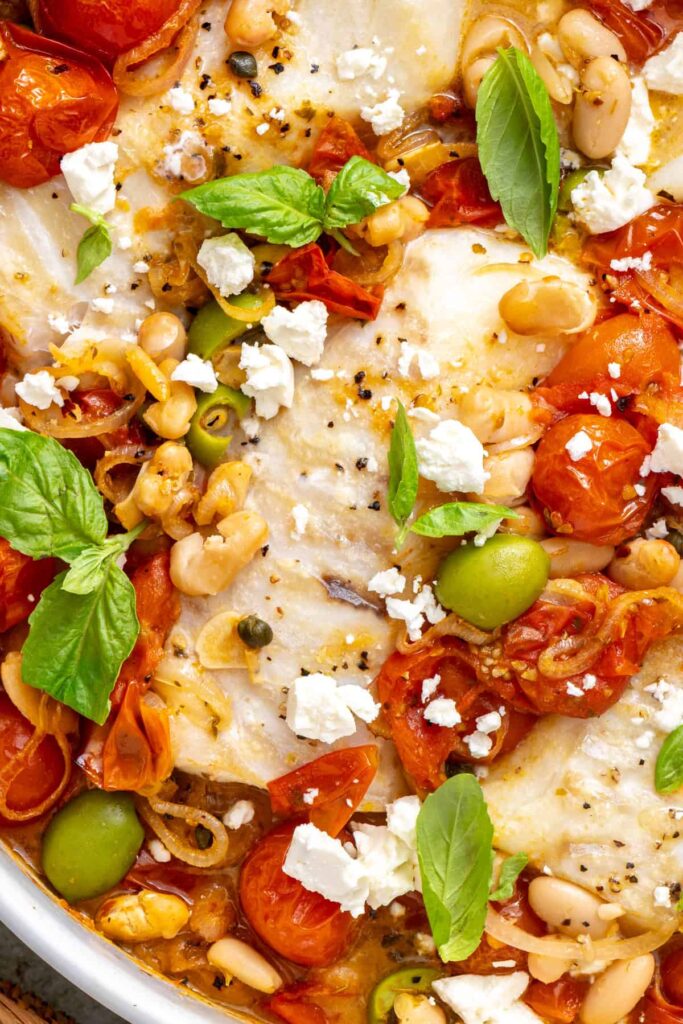 Tight close-up of flaky baked cod with jammy roasted tomatoes, white beans, crumbled feta, and fresh basil.