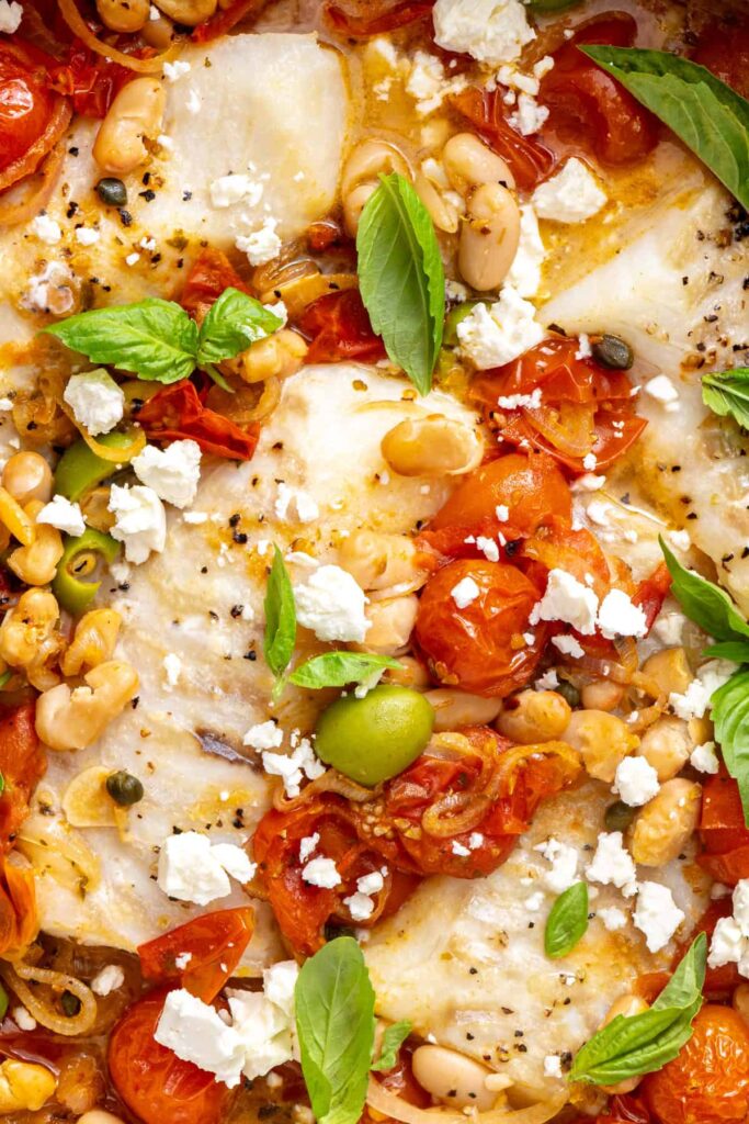 Extreme close-up of flaky baked cod with jammy roasted tomatoes, white beans, crumbled feta, and fresh basil.
