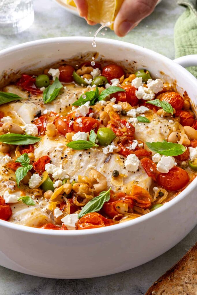 Close-up of lemon juice dripping onto flaky baked cod and roasted tomatoes in a Mediterranean-style fish dish.
