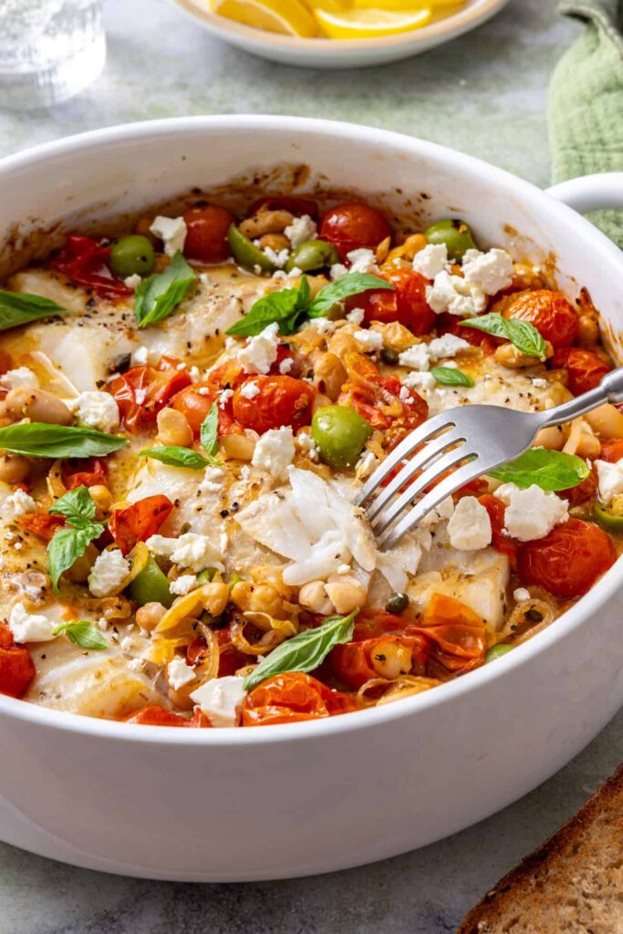 Fork breaking into tender baked cod in a skillet of roasted cherry tomatoes, white beans, feta, and fresh basil.