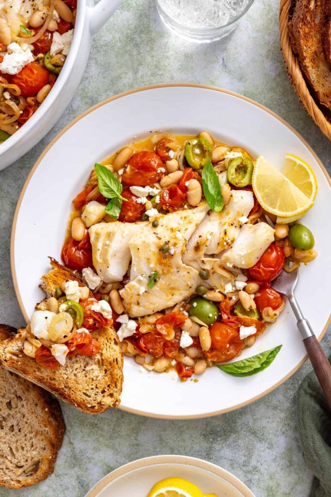 Mediterranean cod with fresh basil and crumbled feta served on a plate with toasted bread for dipping into the tomato sauce.