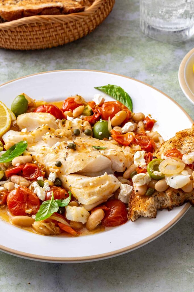 Toasted sourdough topped with flaky baked cod, roasted tomatoes, and white beans, served alongside a plate of Mediterranean cod.