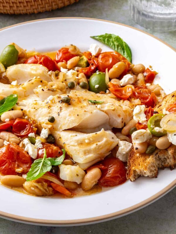 Close-up of Mediterranean baked cod with roasted tomatoes, white beans, feta, and fresh basil, served with sourdough on a plate.