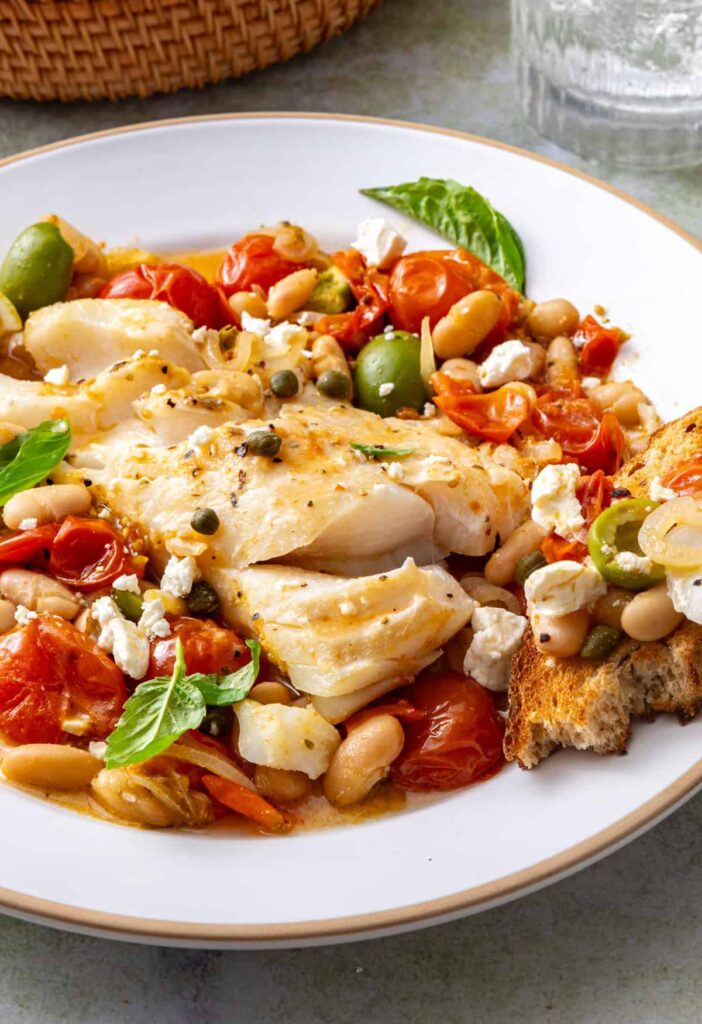 Close-up of Mediterranean baked cod with roasted tomatoes, white beans, feta, and fresh basil, served with sourdough on a plate.