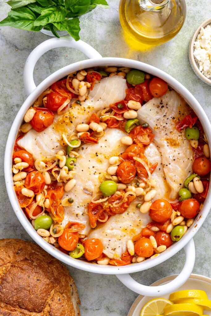 Seasoned cod fillets nestled into a skillet of roasted cherry tomatoes, white beans, olives, and capers before baking.