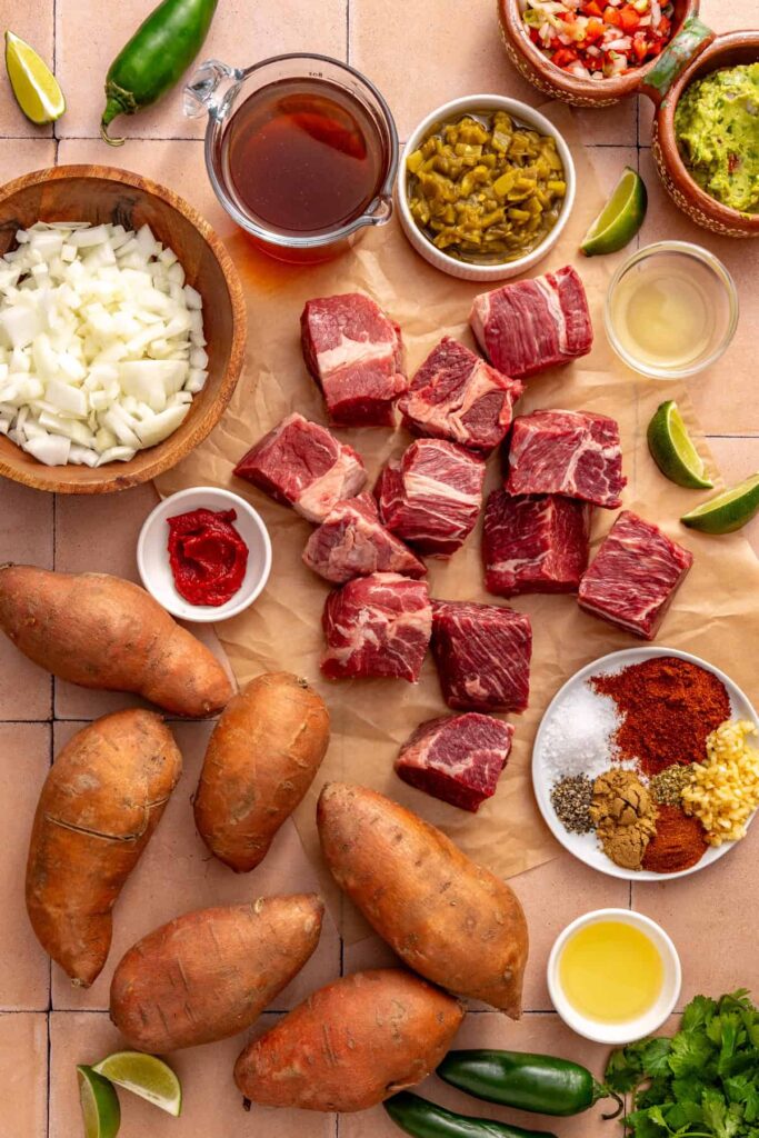 Overhead view of ingredients for Instant Pot beef taco sweet potatoes, including beef chuck roast, sweet potatoes, onion, garlic, spices, broth, green chiles, and lime.