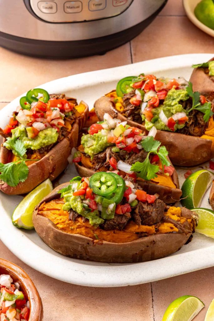Side-angle view of beef taco stuffed sweet potatoes topped with avocado, pico de gallo, cilantro, and jalapeño, with the Instant Pot in the background.