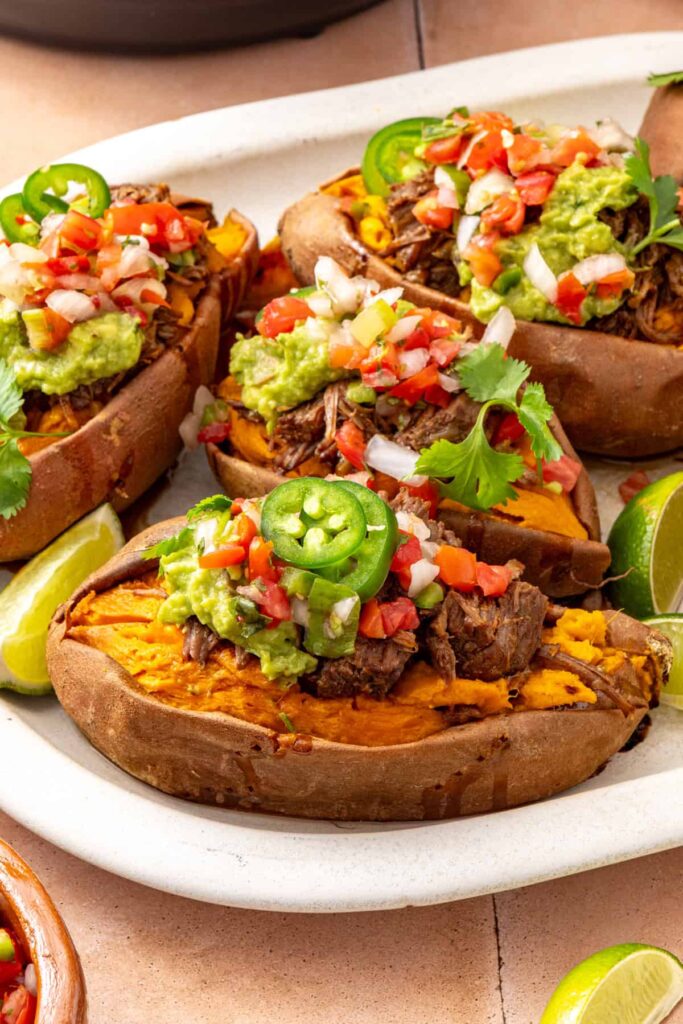 Close side-angle view of beef stuffed sweet potatoes topped with fresh avocado, pico de gallo, and cilantro, with taco toppings nearby.