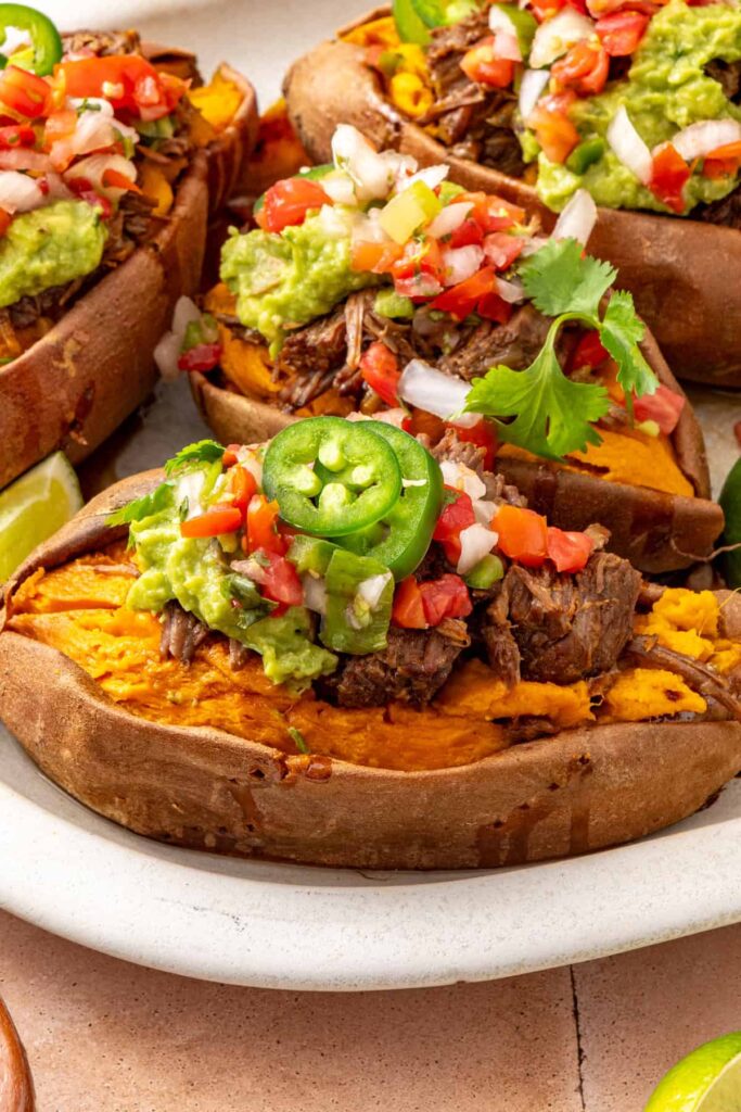 Platter of shredded beef taco sweet potatoes with avocado, cilantro, and lime, styled with small bowls of fresh toppings.