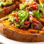 Close-up of an Instant Pot beef taco stuffed sweet potato topped with shredded beef, avocado, pico de gallo, and fresh cilantro, with more stuffed sweet potatoes blurred in the background.