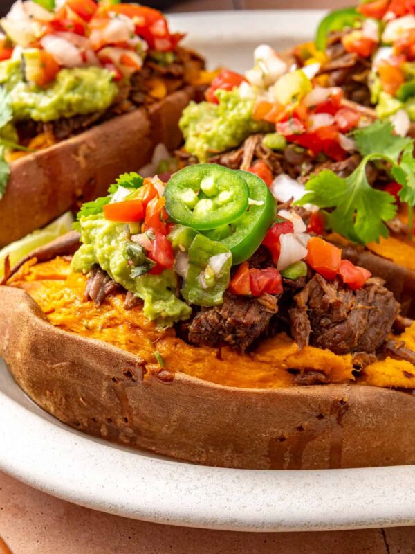 Close-up of an Instant Pot beef taco stuffed sweet potato topped with shredded beef, avocado, pico de gallo, and fresh cilantro, with more stuffed sweet potatoes blurred in the background.