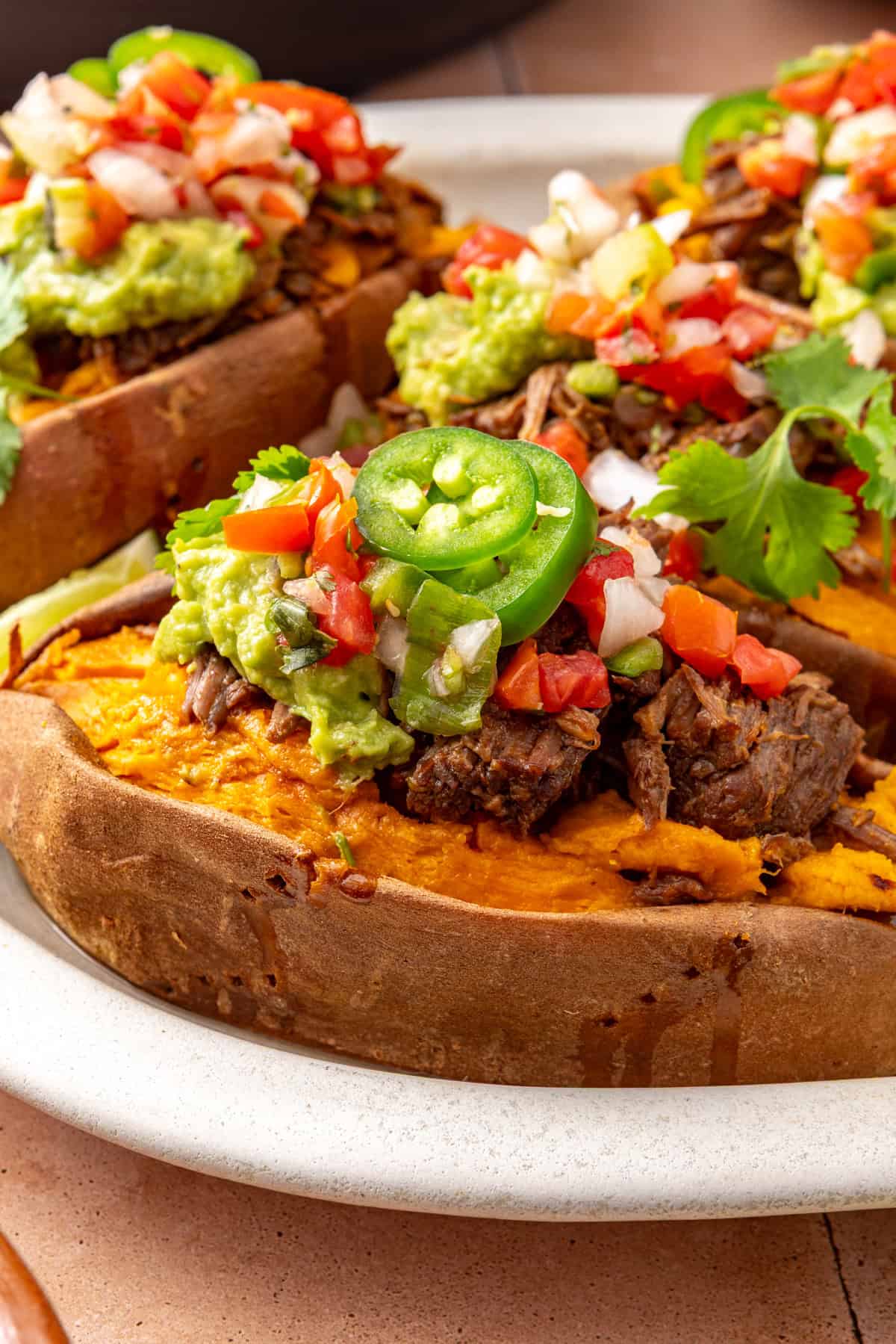 Close-up of an Instant Pot beef taco stuffed sweet potato topped with shredded beef, avocado, pico de gallo, and fresh cilantro, with more stuffed sweet potatoes blurred in the background.