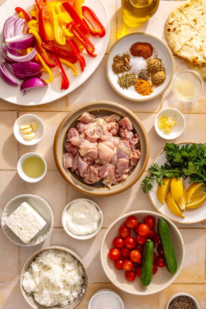 Ingredients for sheet pan chicken shawarma bowls, including chicken thighs, bell peppers, red onion, spices, feta, Greek yogurt, rice, cucumber, tomatoes, lemon, and fresh parsley.