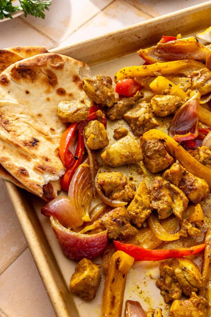 Overhead sheet pan chicken shawarma with roasted bell peppers and red onion, served alongside warm pita bread.