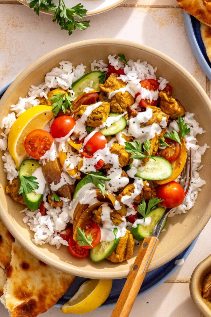 Close-up overhead chicken shawarma bowl featuring roasted chicken, crisp cucumber, juicy tomatoes, and a drizzle of garlic feta sauce.