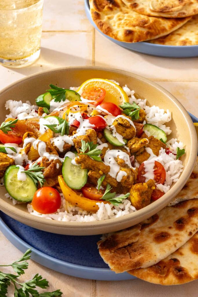Side-angle close-up of a chicken shawarma bowl with basmati rice, roasted chicken and peppers, fresh cucumber, tomatoes, and creamy feta garlic sauce.