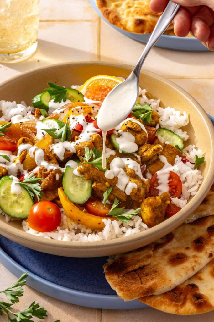 Hand drizzling creamy feta garlic sauce over a chicken shawarma bowl with roasted chicken, basmati rice, and fresh vegetables.