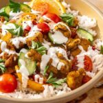 Side-angle close-up of a chicken shawarma bowl with roasted chicken, colorful vegetables, creamy feta garlic sauce, fresh parsley, and pita bread with a lemon wedge on the side.