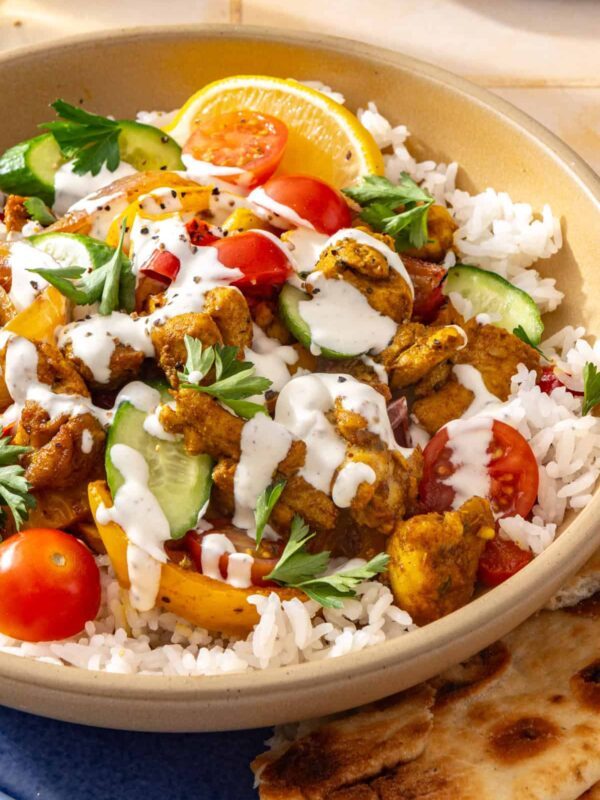 Side-angle close-up of a chicken shawarma bowl with roasted chicken, colorful vegetables, creamy feta garlic sauce, fresh parsley, and pita bread with a lemon wedge on the side.