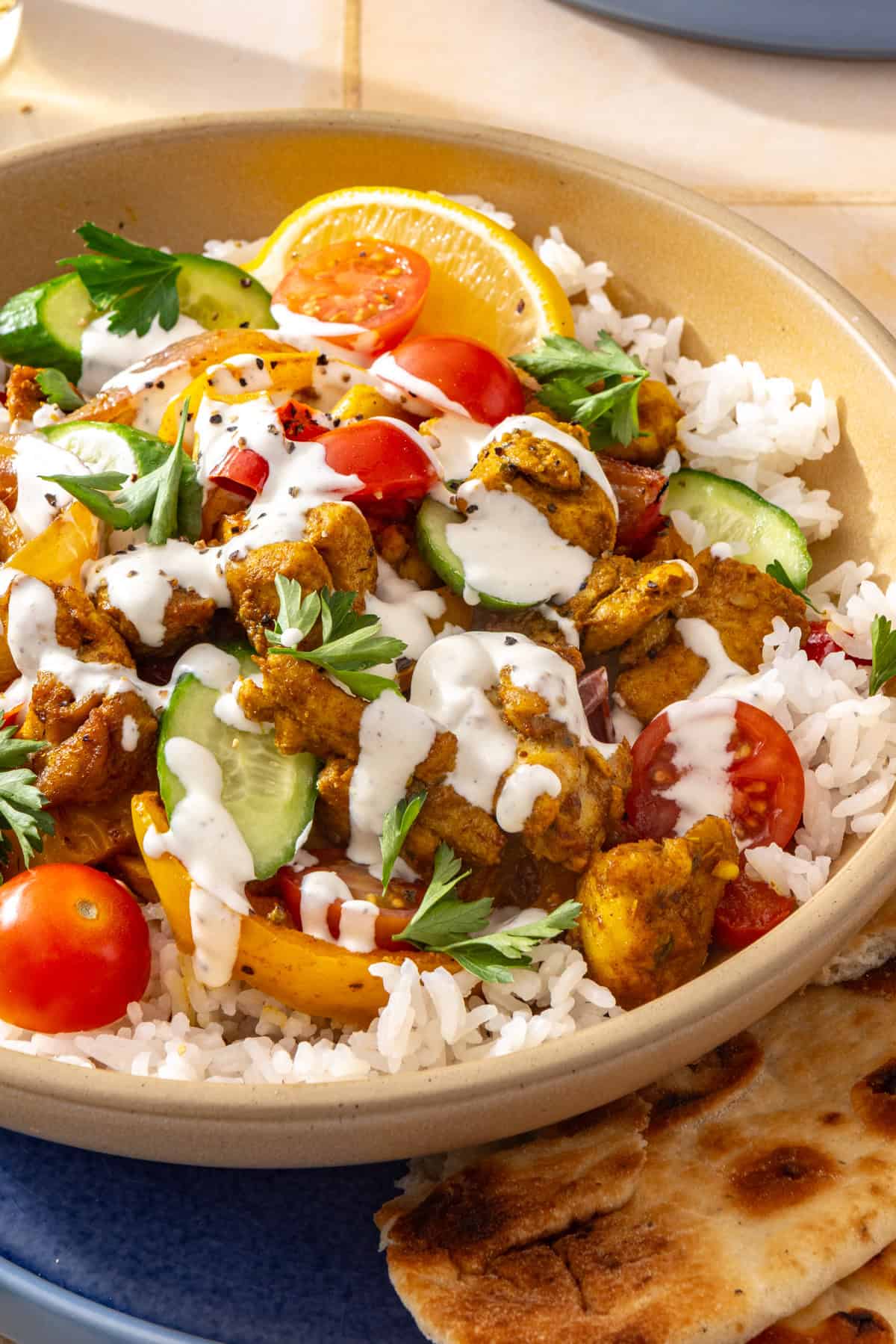 Side-angle close-up of a chicken shawarma bowl with roasted chicken, colorful vegetables, creamy feta garlic sauce, fresh parsley, and pita bread with a lemon wedge on the side.